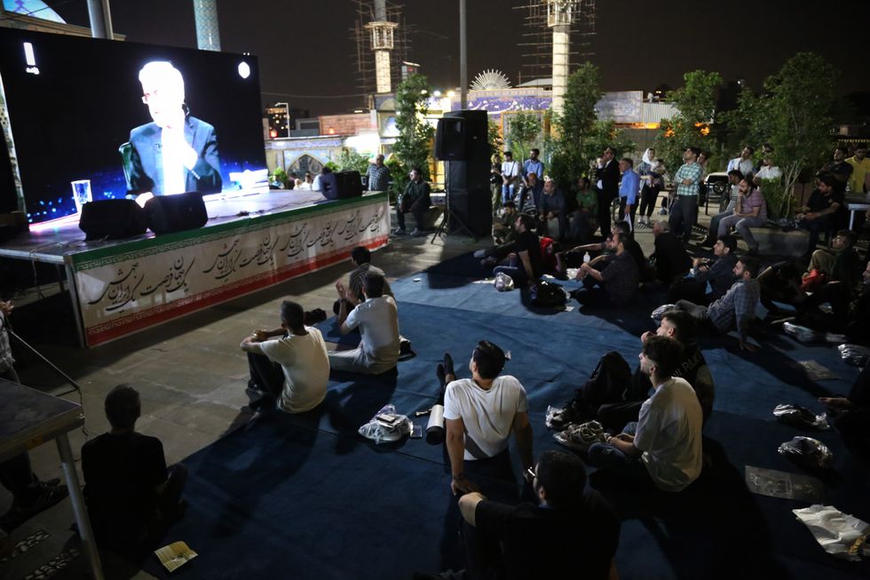 Photo of a community gathered around to look at a giant screen on a stage showing the video of a speech