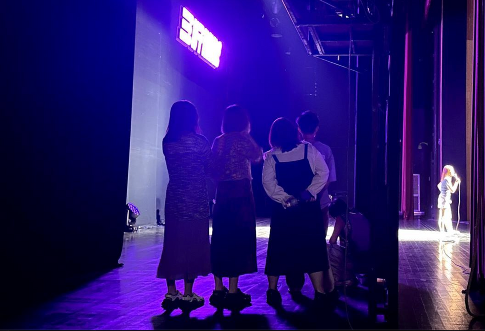 Photo of a group of comedians waiting in the wings as a female stand-up comedian performs on stage at Beijing's ​Laughing Fruit Factory