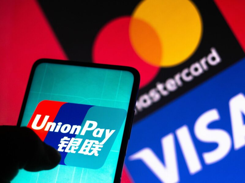 Photo of a hand holding a phone displaying an Union Pay logo, with a Mastercard VISA logo in the background of the photo.
