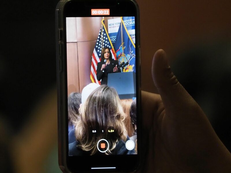 Photo of a hand holding a phone recording footage of ​Vice President Kamala Harris during a midterm rally in NYC on Nov. 3