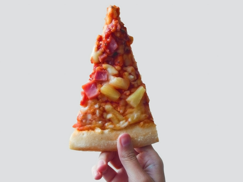Photo of a hand holding a slice of Hawaiian pizza