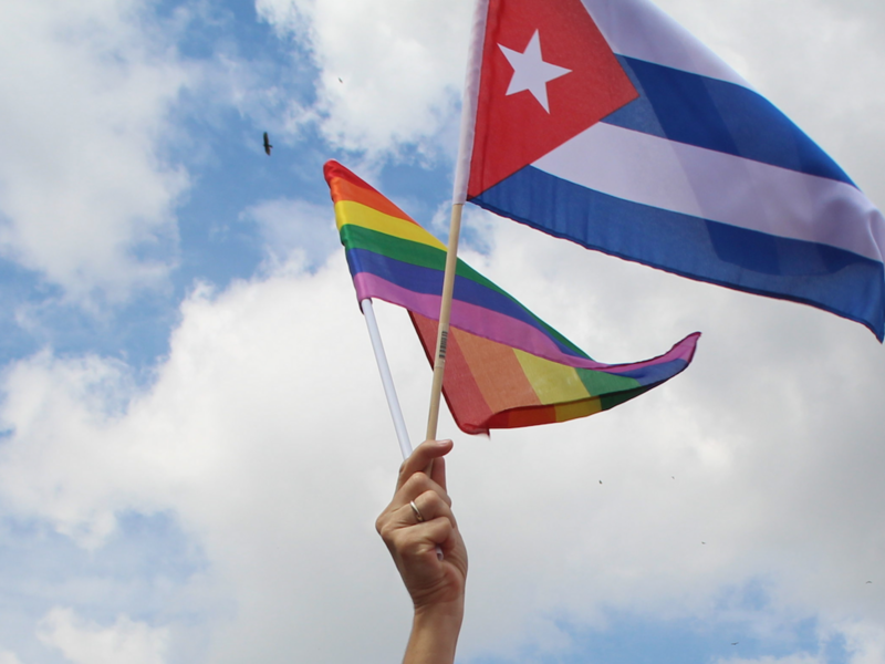 Photo of a hand waving a rainbow flag and a Cuba flag at a pro-LGBTQ+ rights demonstration in Havana, Cuba