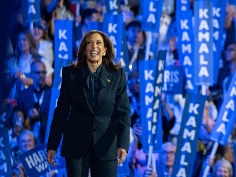Photo of a Kamala Harris walking onstage at a rally, with a crowd of people waving blue signs reading "Kamala" in the background