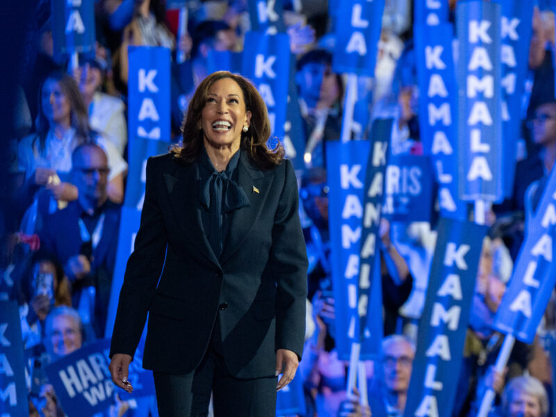 Photo of a Kamala Harris walking onstage at a rally, with a crowd of people waving blue signs reading "Kamala" in the background