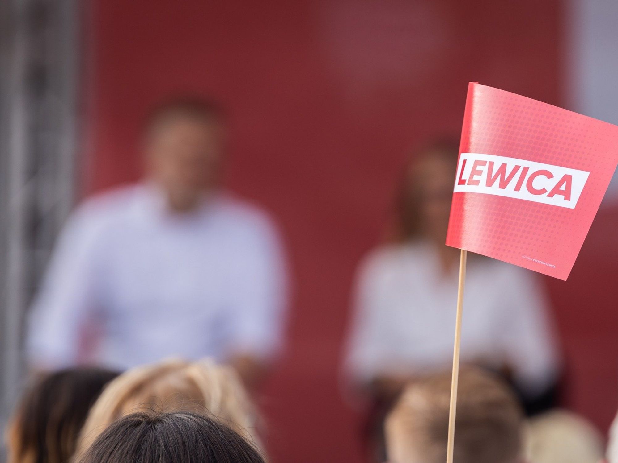 Photo of a Lewica ("Left") meeting in Warsaw, Poland, with a flag from the left-wing party in focus while members of the crowd and participants are out of focus