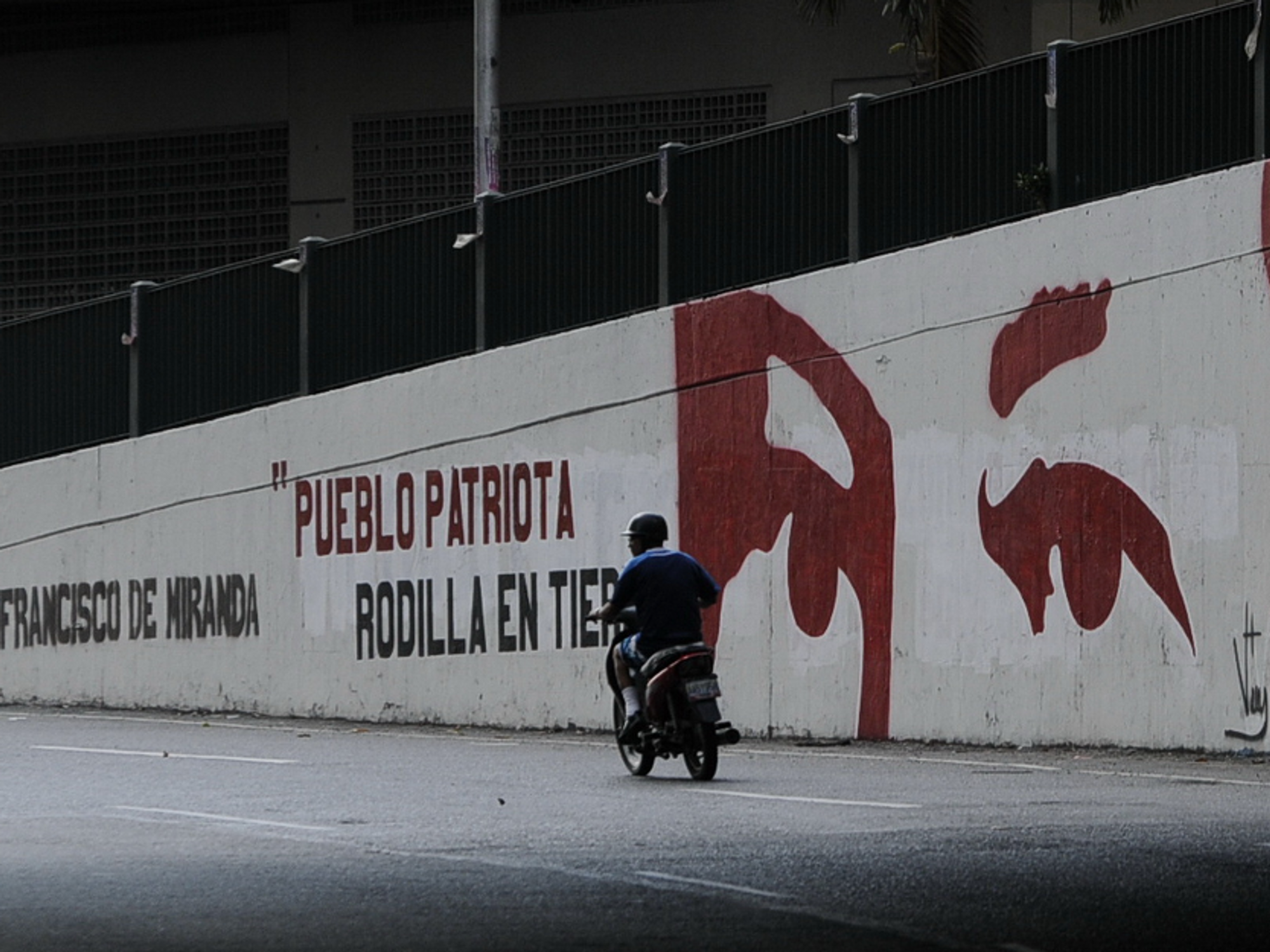Photo of a man driving a motorbike past a wall with a mural depicting former President Hugo Chavez in Caracas, Venezuela