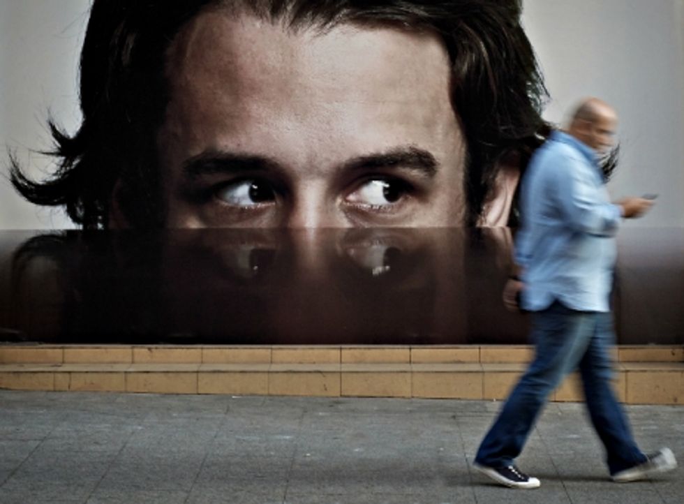 Photo of a man on his phone walking past a billboard of a big face watching him