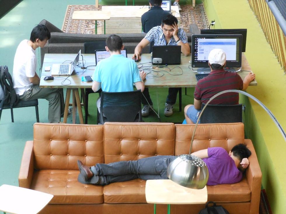 photo of a man sleeping on a couch while others work on their computers.