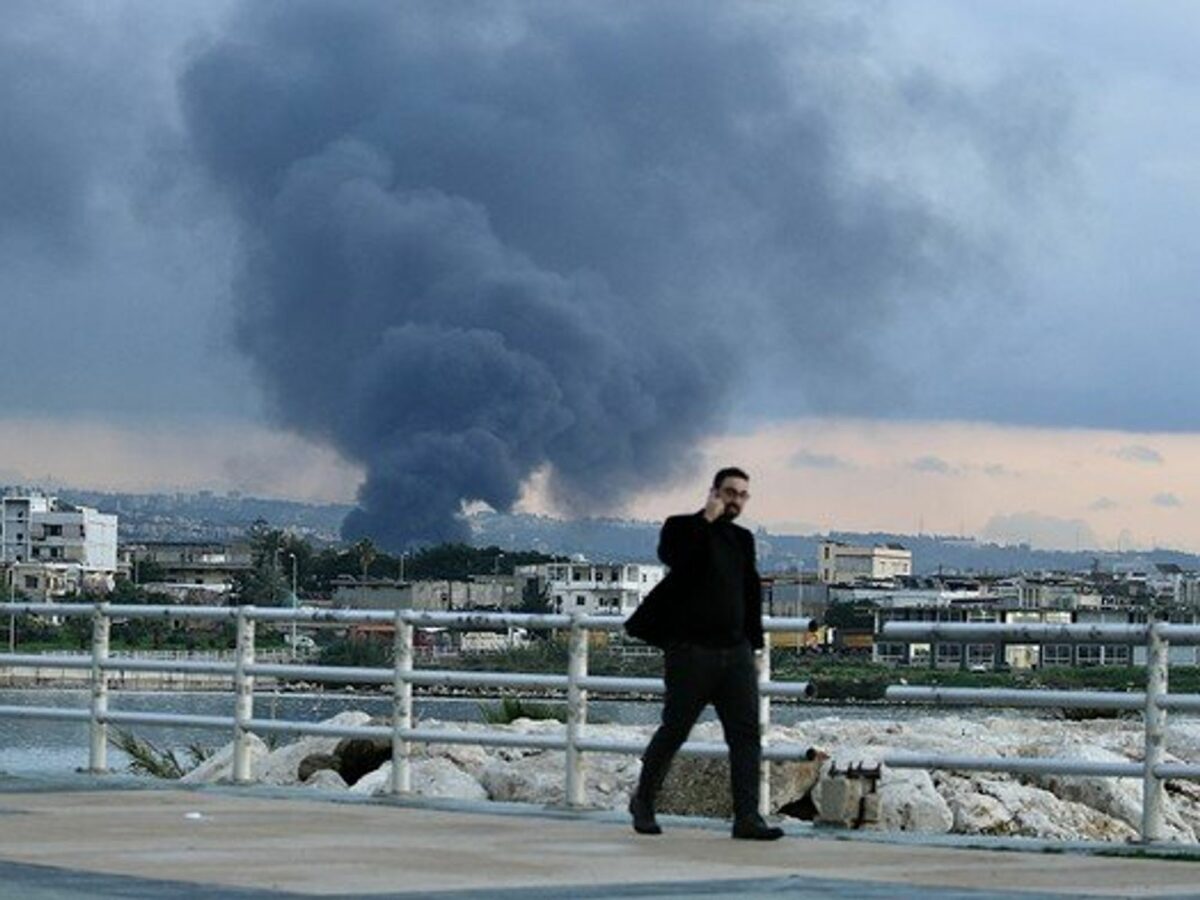 Photo of a man walking in Ghazieh, Lebanon, as plumes of smoke rise in the background