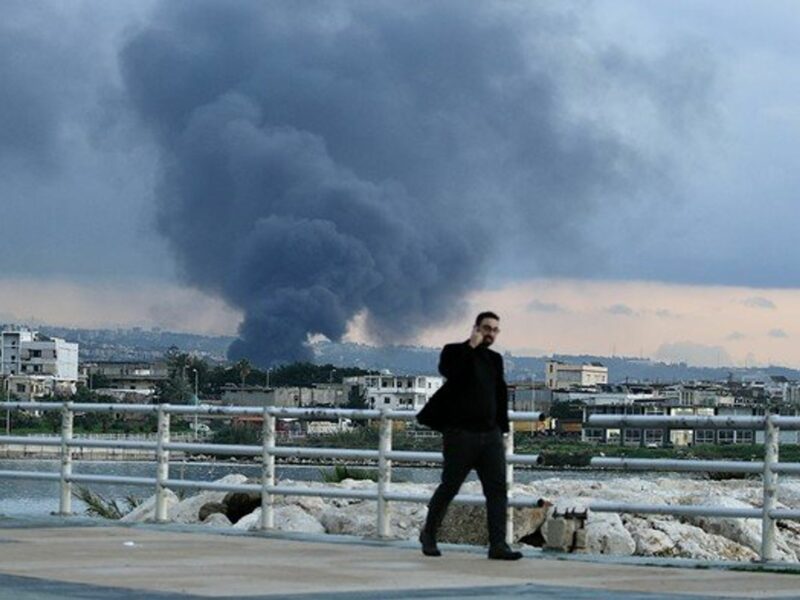Photo of a man walking in Ghazieh, Lebanon, as plumes of smoke rise in the background