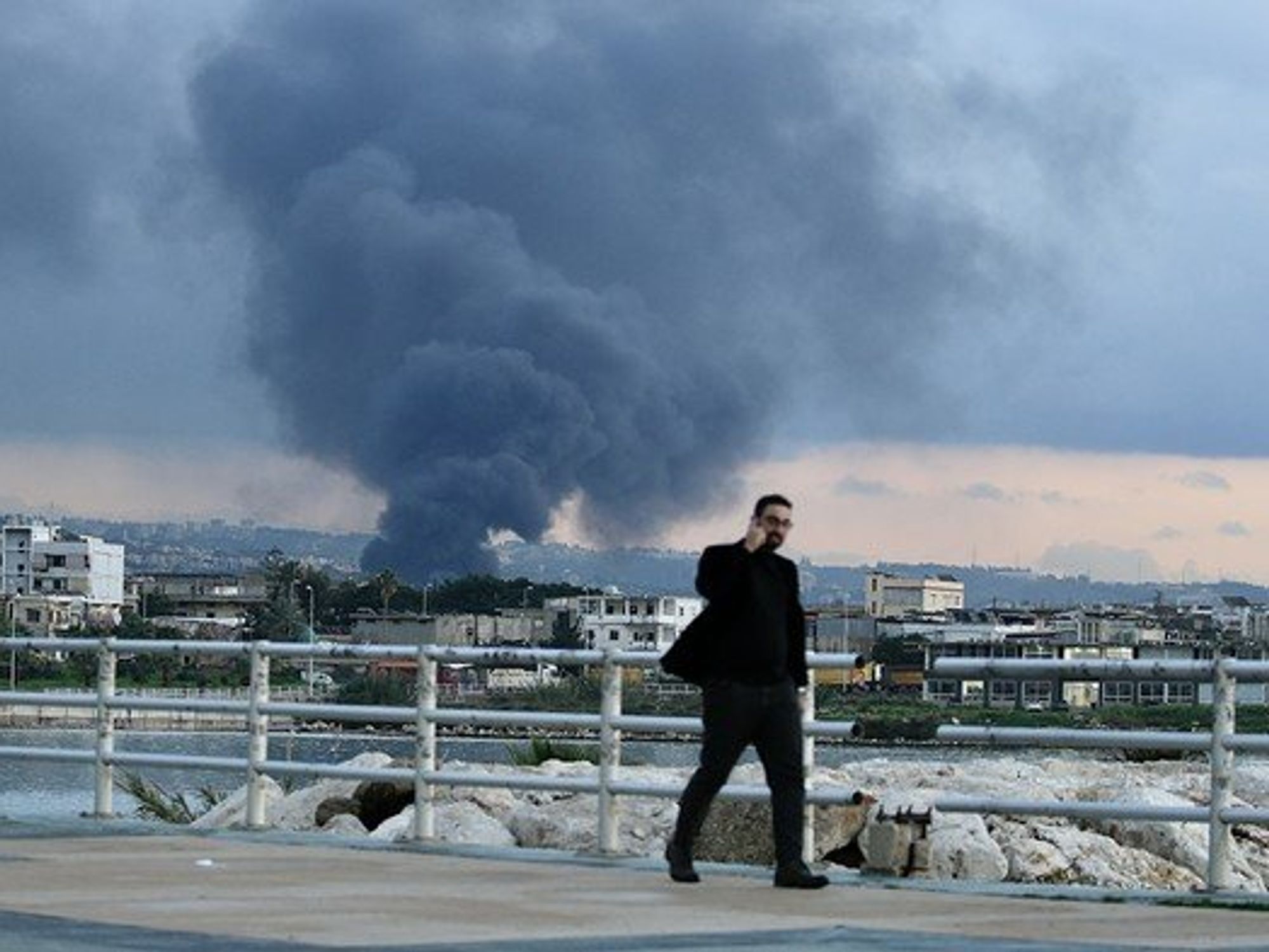 Photo of a man walking in Ghazieh, Lebanon, as plumes of smoke rise in the background