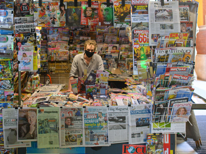 Photo of a man wearing a black COVID-19 protective mask inside a newsstand, surrounded by dozens of newspapers and magazines, in Budapest, Hungary.