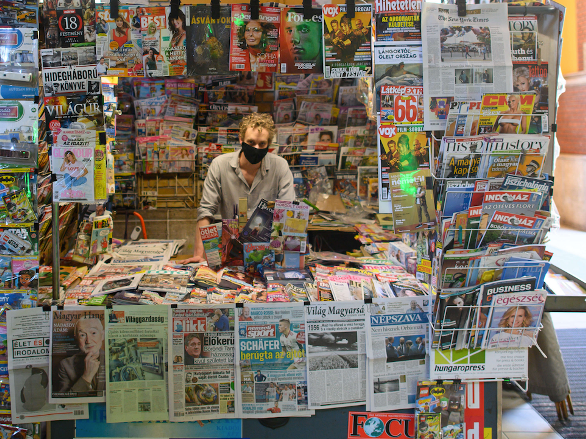 Photo of a man wearing a black COVID-19 protective mask inside a newsstand, surrounded by dozens of newspapers and magazines, in Budapest, Hungary.
