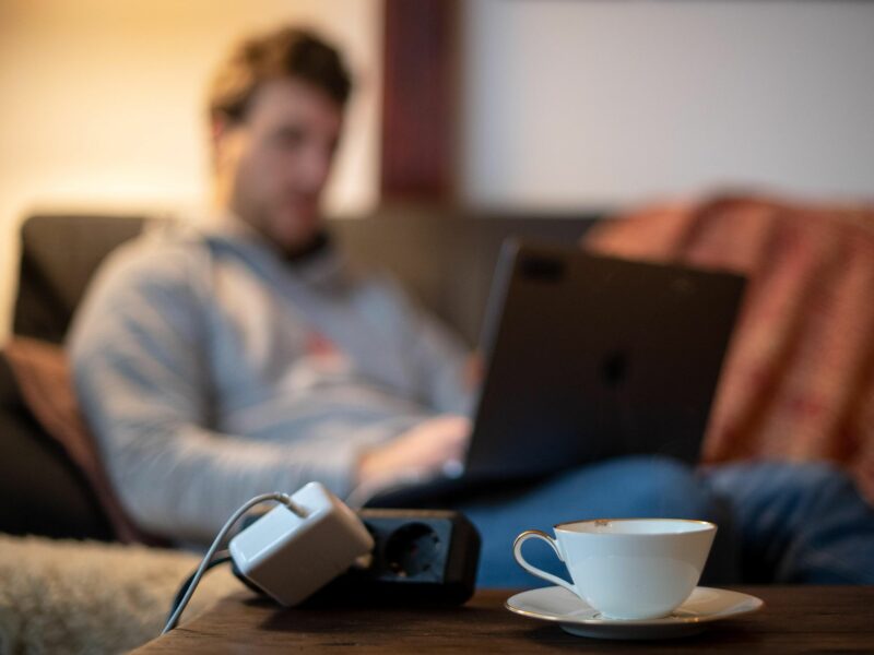 Photo of a man working on his laptop while sat on a couch, with a power plug and a cup of tea in the foreground.