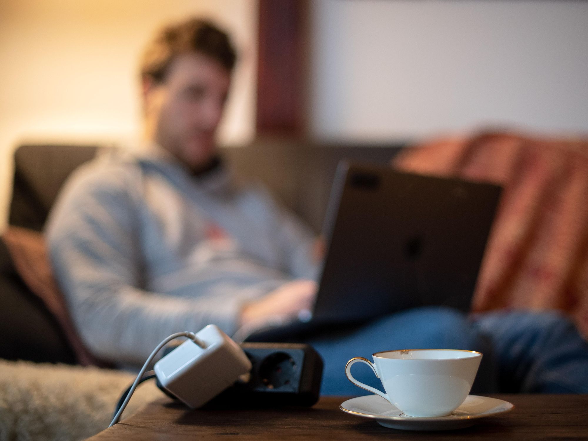 Photo of a man working on his laptop while sat on a couch, with a power plug and a cup of tea in the foreground.