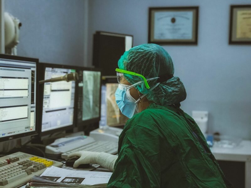 Photo of a medical worker covered in green protective gear looking at a computer screen