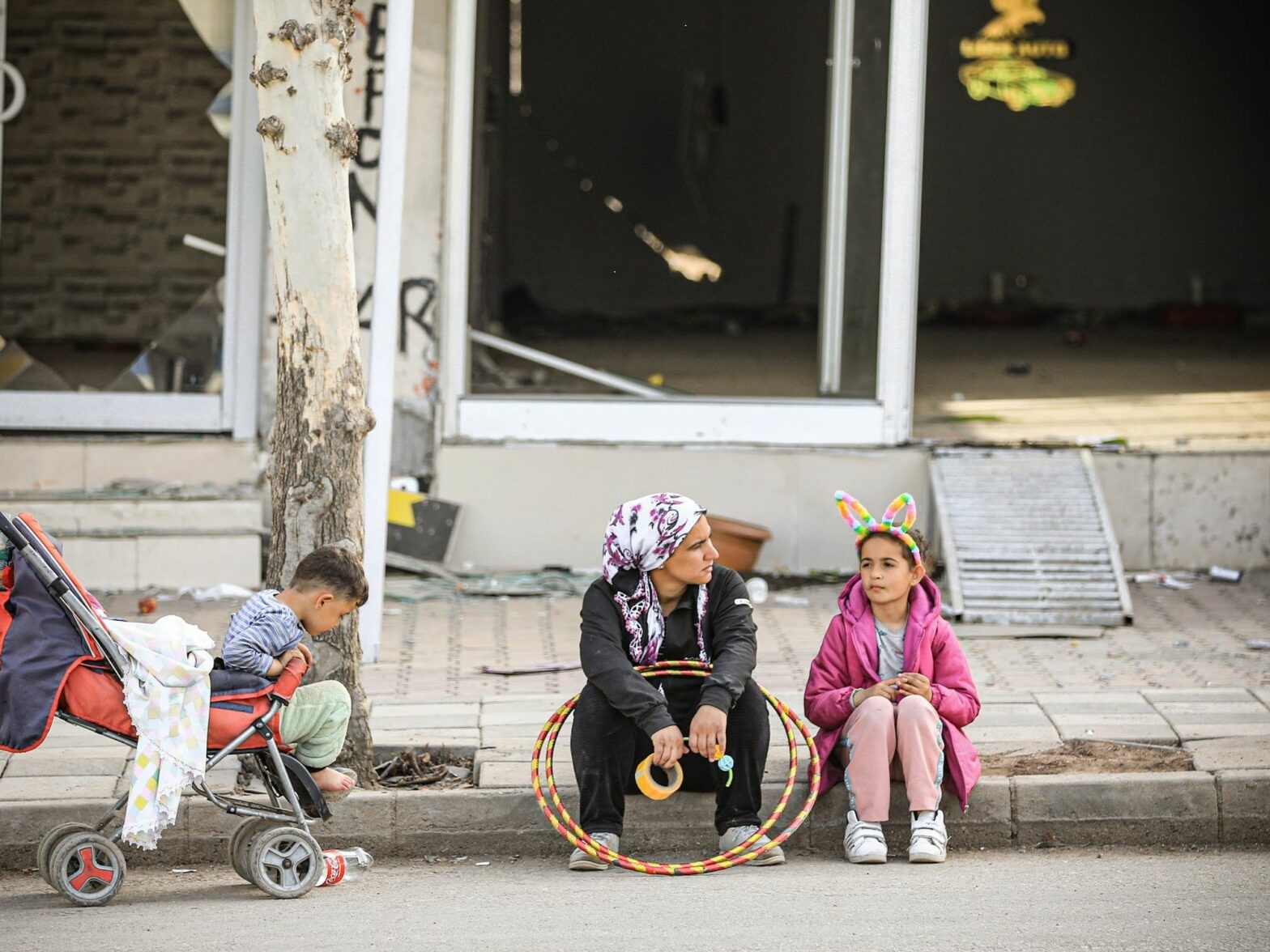 Photo of a mother and two children sitting on the sidewalk.