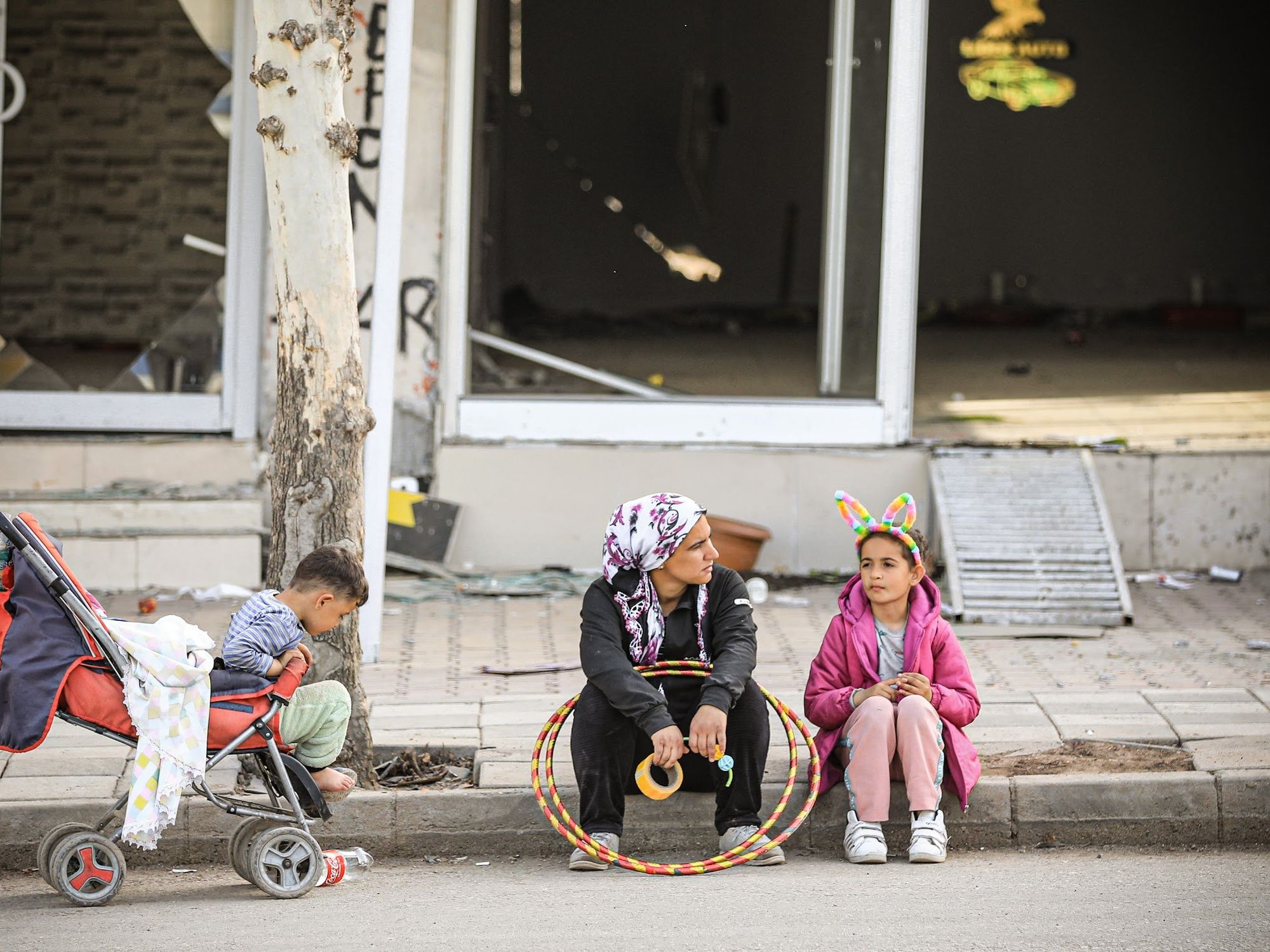 Photo of a mother and two children sitting on the sidewalk.
