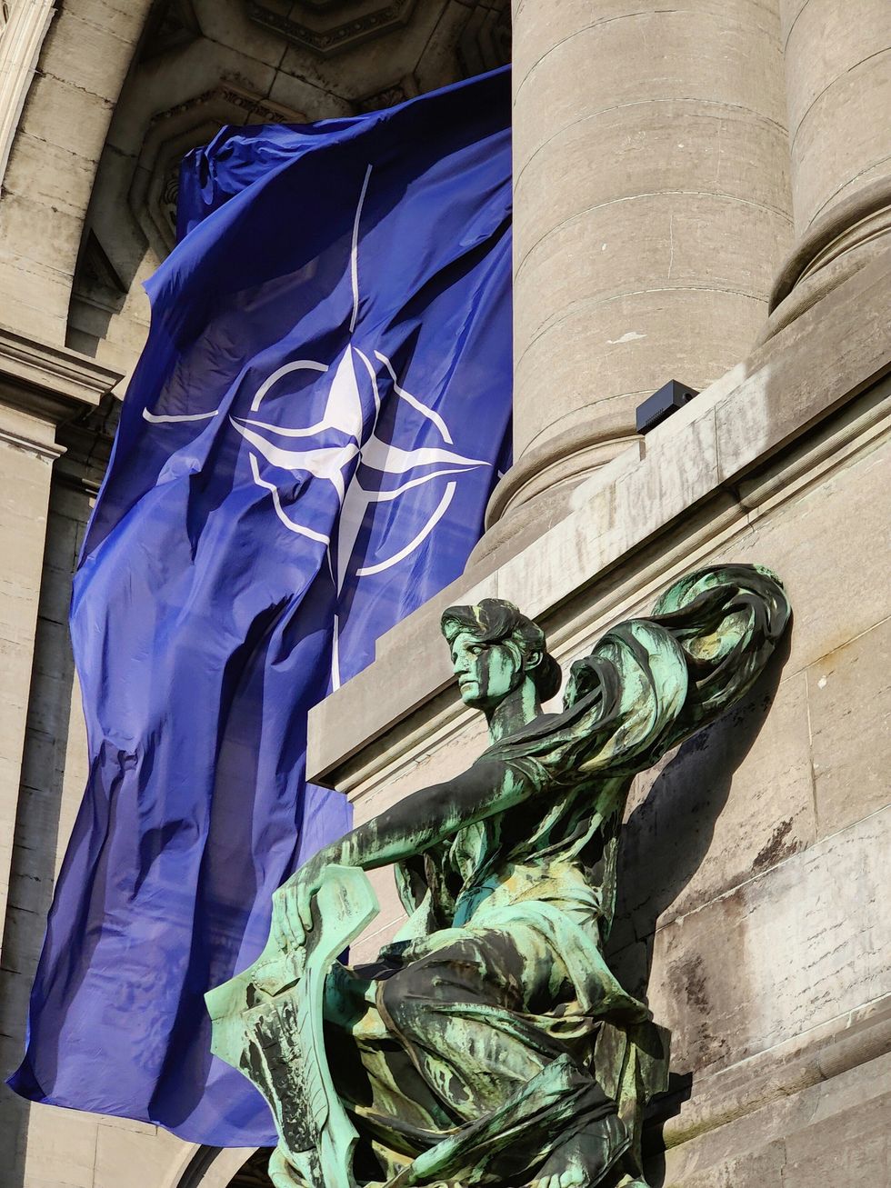 Photo of a NATO flag floating in Brussels, Belgium, behind a bronze statue.