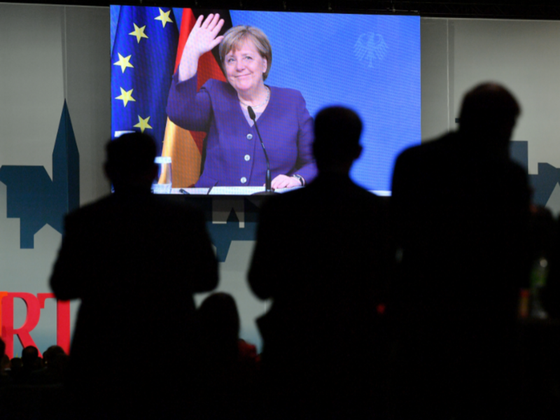 Photo of a people in front of a TV screen displaying Chancellor Angela Merkel waving