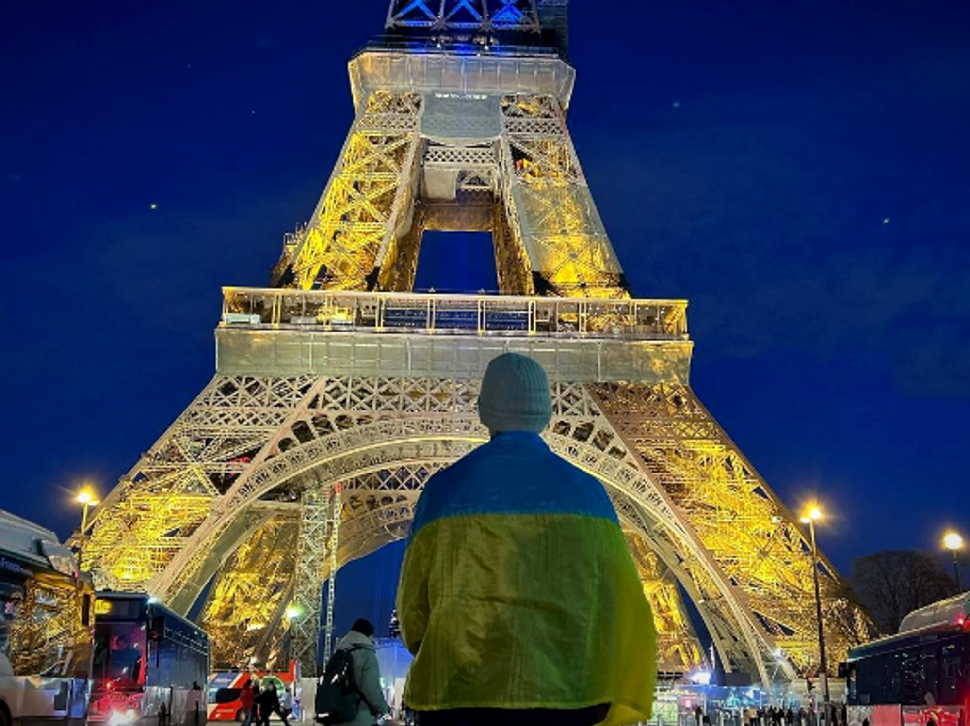 Photo of a person draped in a Ukraine flag standing in front of the Eiffel Tower, lit up in yellow and blue to mark the one-year anniversary of the war in Ukraine