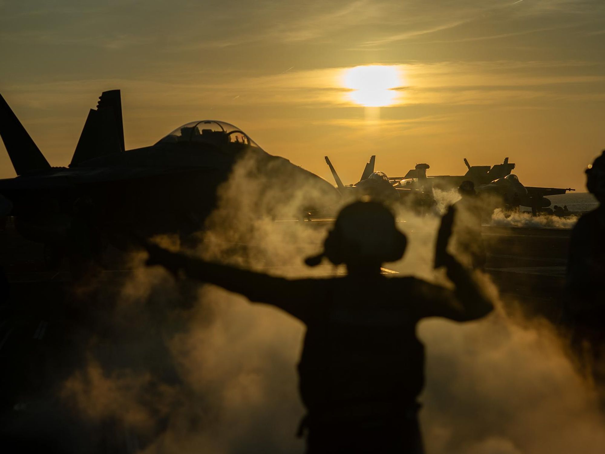 Photo of a sunset over the USS Nimitz with a man guiding fighter jets ready for takeoff
