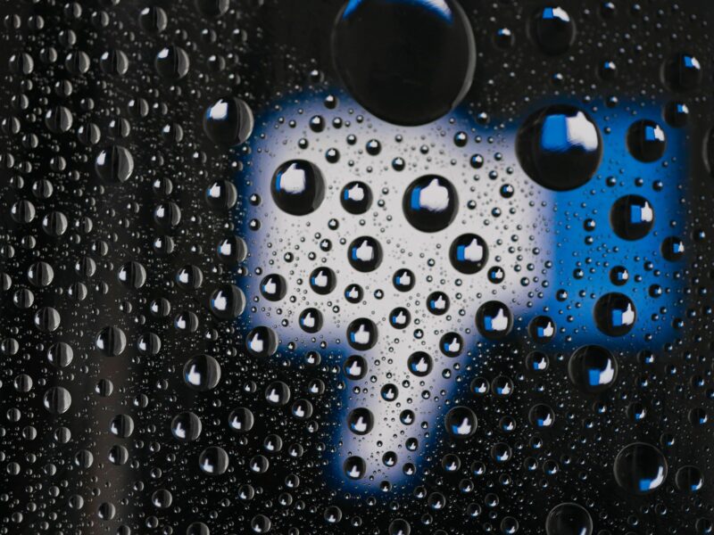 Photo of a window pane with water droplets reflecting Facebook's thumb up logo, with one big thumb down in the background