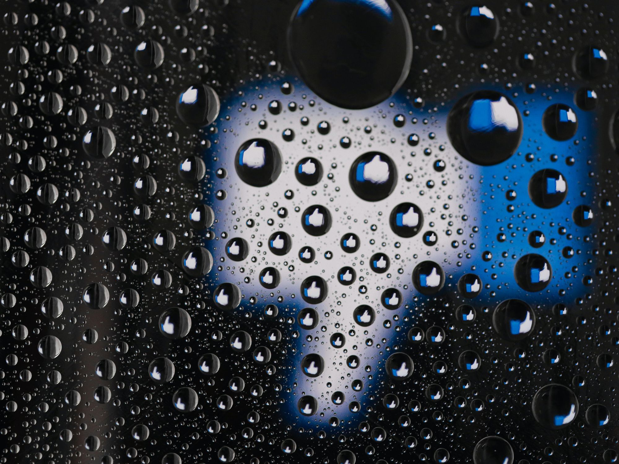 Photo of a window pane with water droplets reflecting Facebook's thumb up logo, with one big thumb down in the background