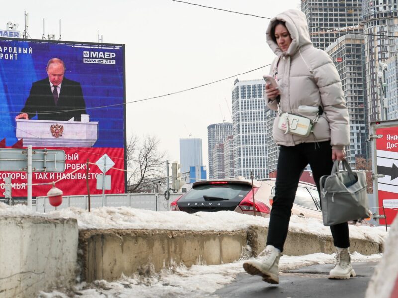 Photo of a woman checking her phone as she walks past a screen showing a broadcast of President Vladimir Putin speaking