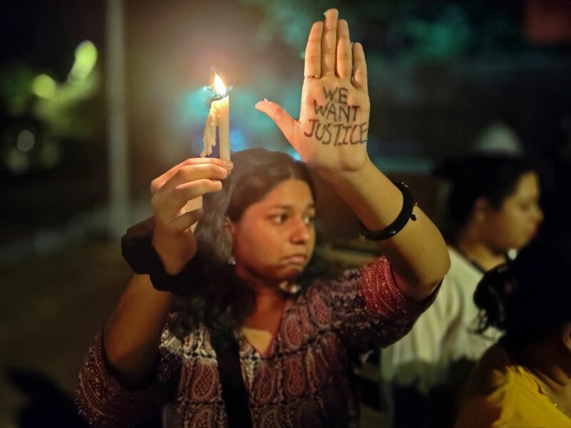 Photo of a woman holding a candle in one hand and raising her other hand, on which the words "we want justice" are written, during a protest in Mumbai, India.