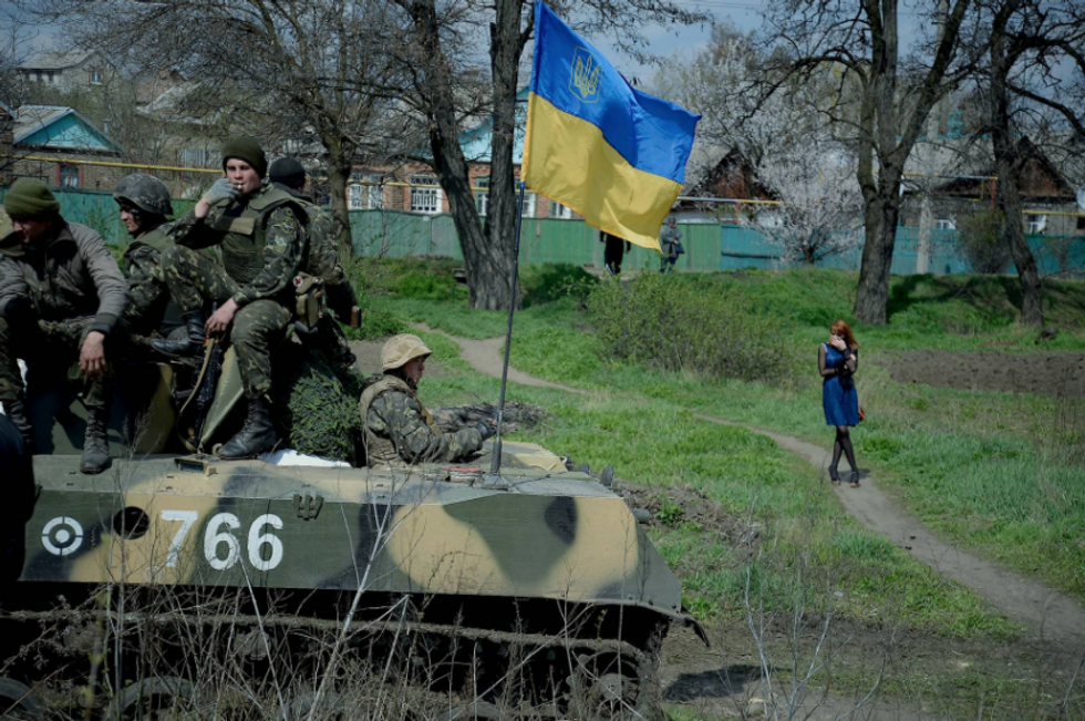 Photo of a woman in a blue dress walking by a tank flying a Ukrainian flag