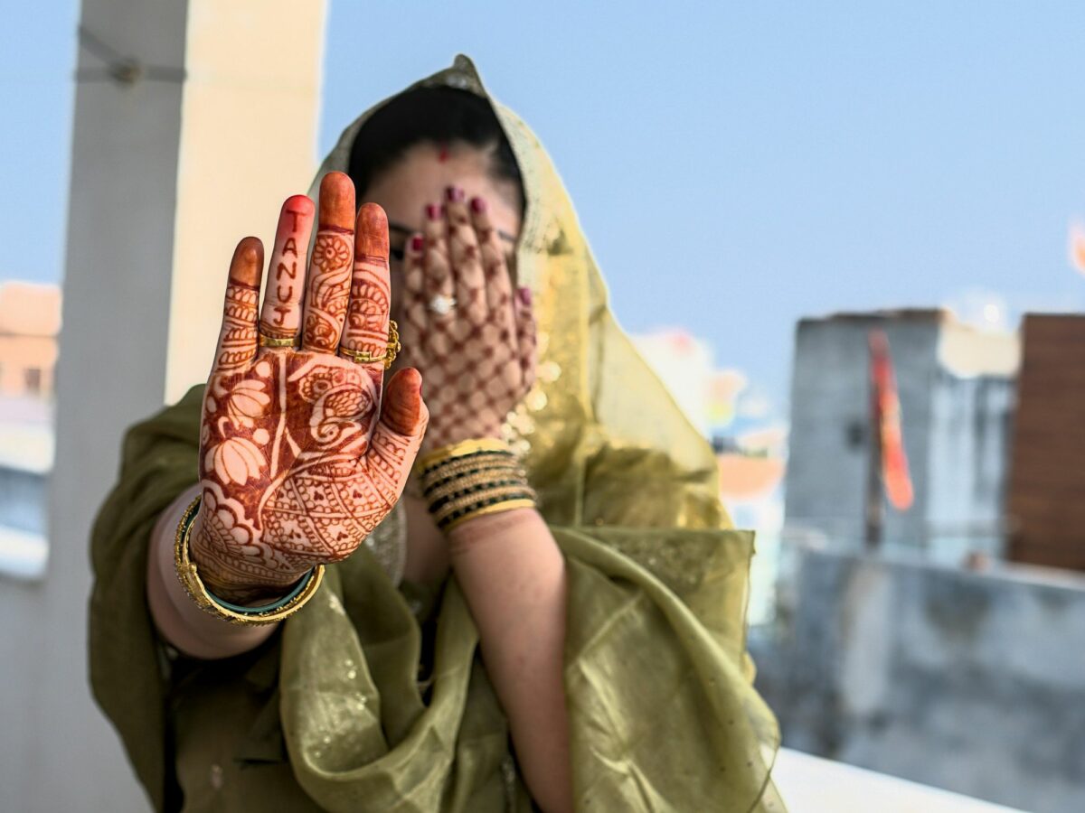 Photo of a woman in a green sari holding one hand up to her face and showing her other, henna-tattooed hand to the camera.