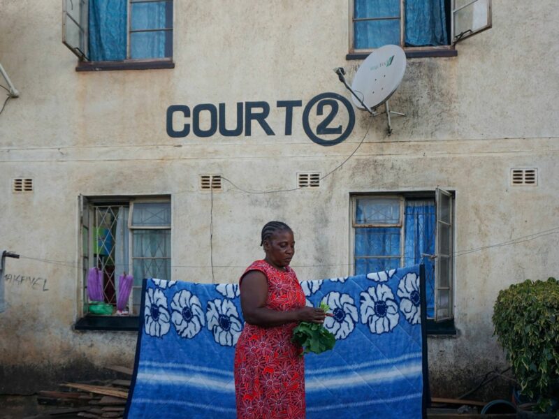 Photo of a woman standing outside her house in Mutare, Zimbabwe, holding leaves in her hand