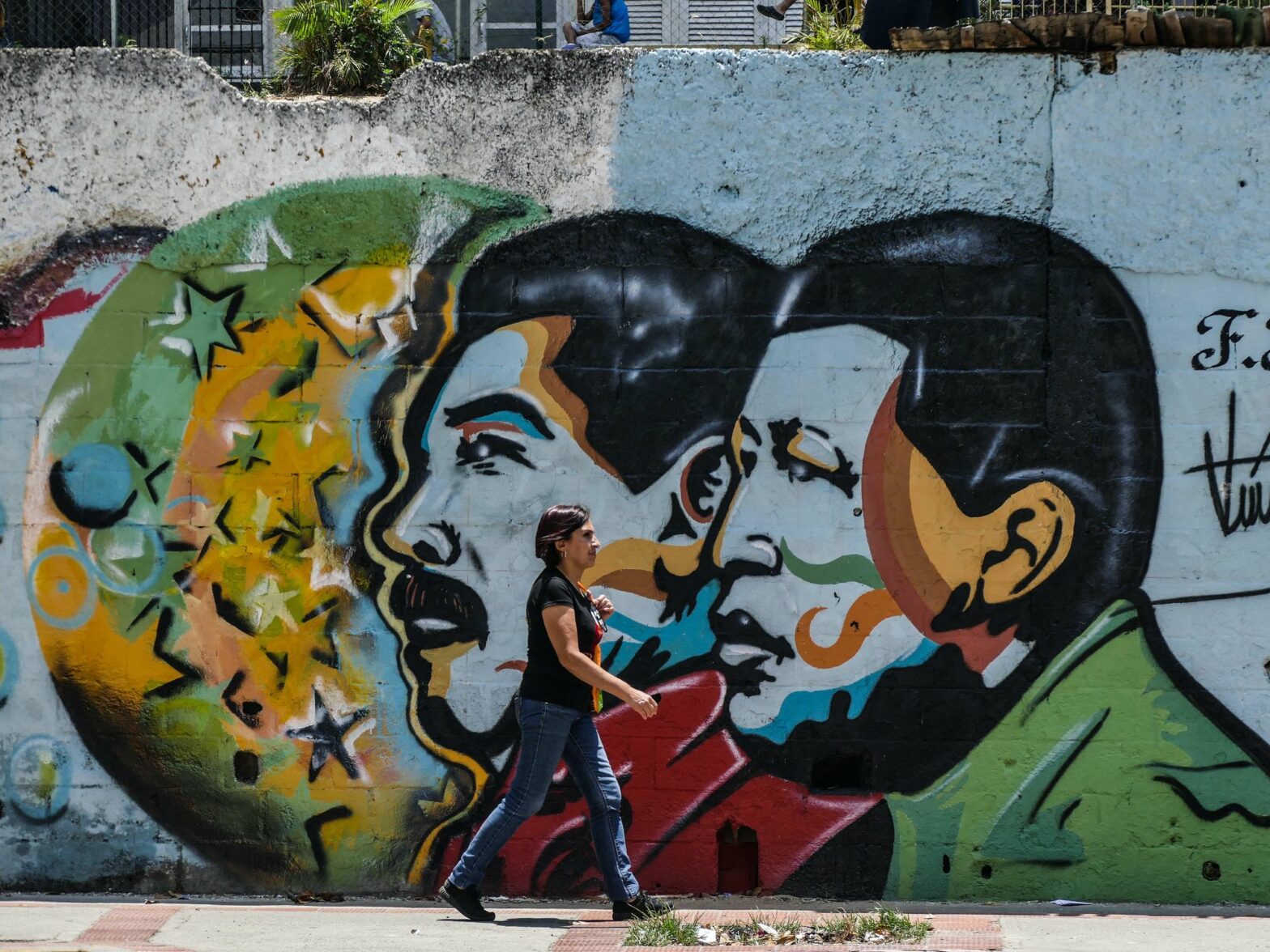 Photo of a woman walking by a wall with street art of Venezuelan presidents Maduro and Chavez in Caracas
