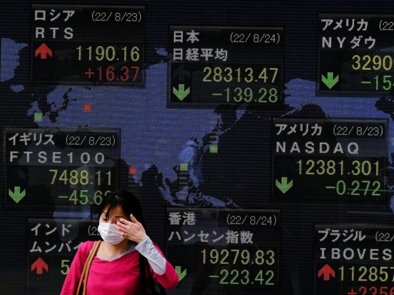 Photo of a woman walking in front of a panel showing downward trends at the Tokyo stock exchange