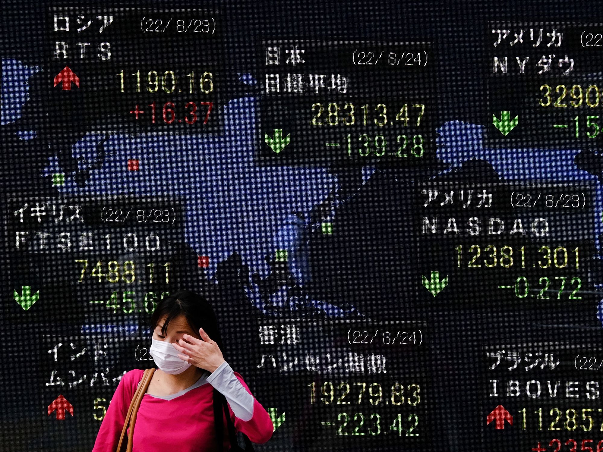 Photo of a woman walking in front of a panel showing downward trends at the Tokyo stock exchange