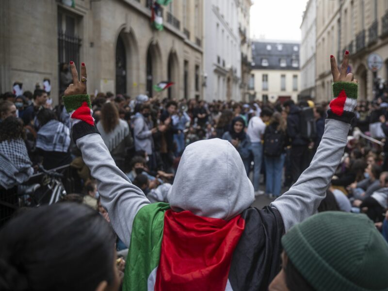 Photo of a woman wearing a Palestinian flag on her shoulders, raising her arms while doing V signs with her hands, as part of a pro-Palestinian protest in Paris