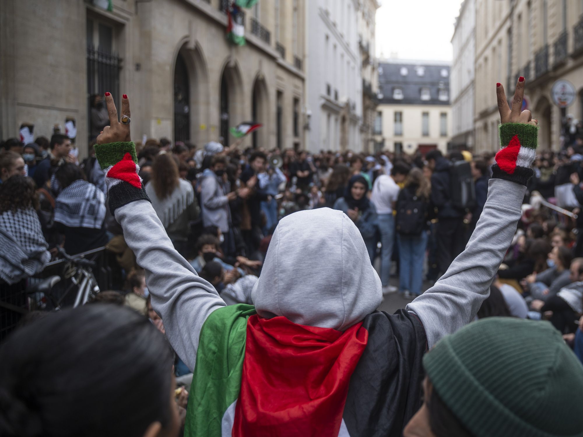 Photo of a woman wearing a Palestinian flag on her shoulders, raising her arms while doing V signs with her hands, as part of a pro-Palestinian protest in Paris