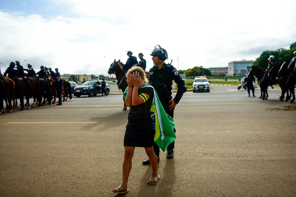 photo of a women supporting former Brazilian President Bolsonaro and wearing a Brazilian flag as a cap is crying as she is escorted out a of camp of Bolsonaro supporters and walks in the middle of policemen on horses