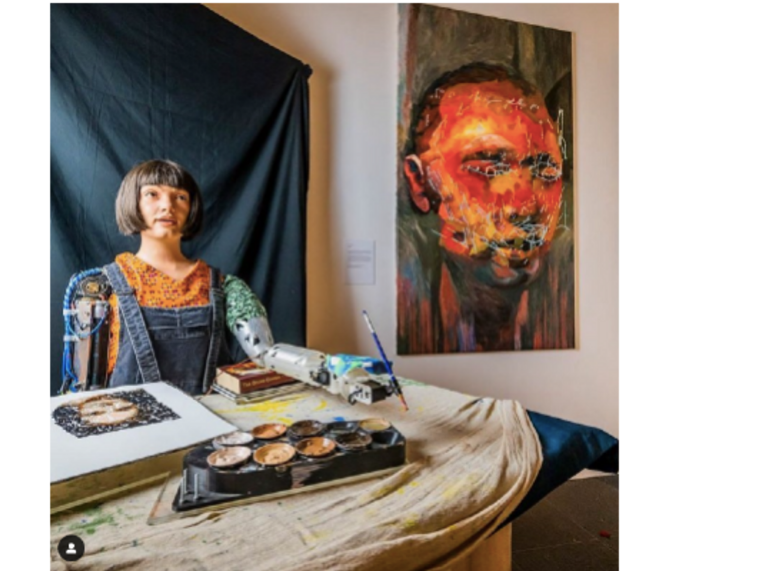 Photo of Ai-Da sitting behind a desk and holding a paint brush