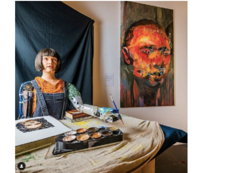 Photo of Ai-Da sitting behind a desk and holding a paint brush