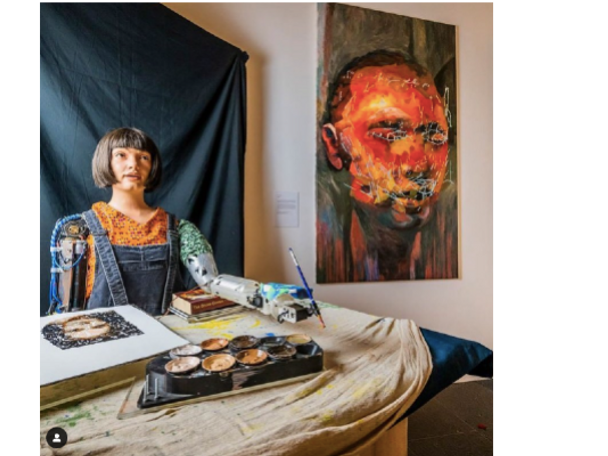 Photo of Ai-Da sitting behind a desk and holding a paint brush