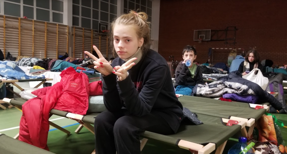 Photo of ​Sofiia sitting on a stretcher bed at a refugee camp, doing V signs with her hands. In the background, her brother Demian is drinking water from a bottle.