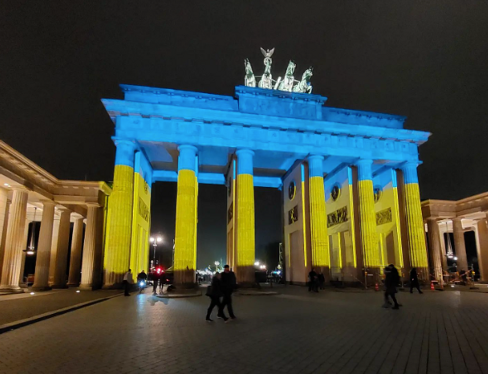 Photo of people walking by ​Berlin's Brandenburg Gate lit up in blue and yellow to mark the one-year anniversary of the Ukraine war