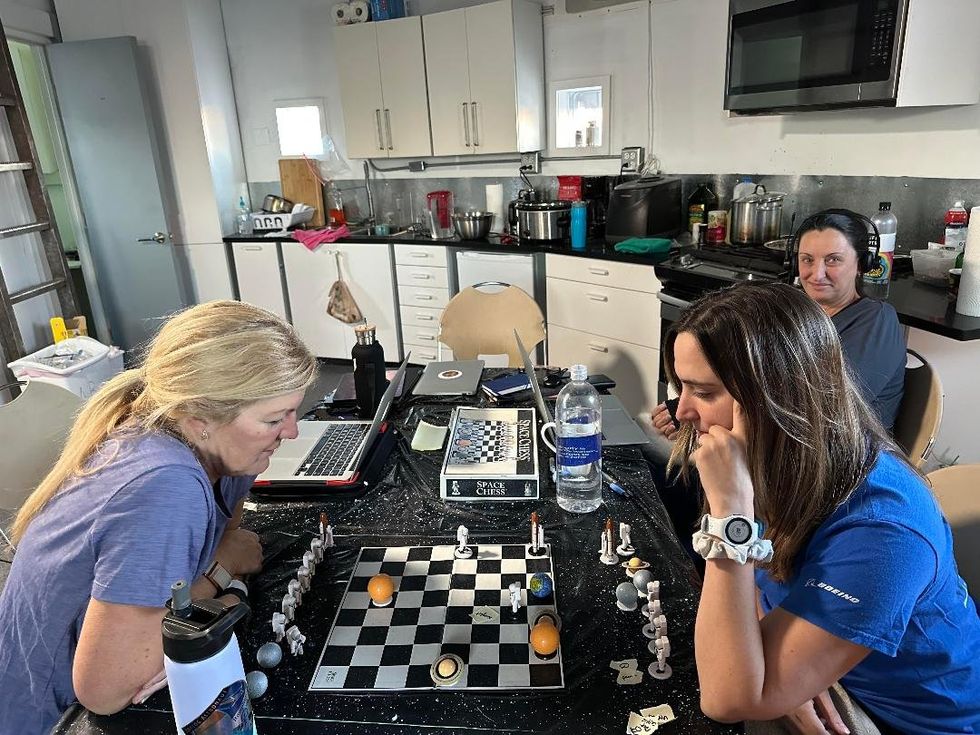 Photo of three women sat in the kitchen of he Mars Desert Research Station, two of whom are playing space-themed chess
