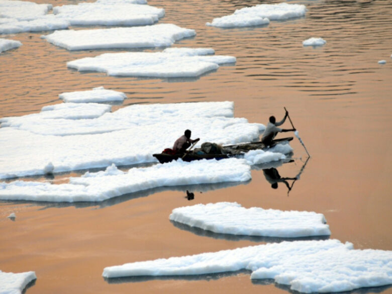 Photo of two fishermen cross through a layer of toxic foam floating on the sacred Yamuna River in India. According to the city government, the foam has been generated by critically high levels of air pollution, untreated sewage water, and industrial waste discharged into the already very polluted river.