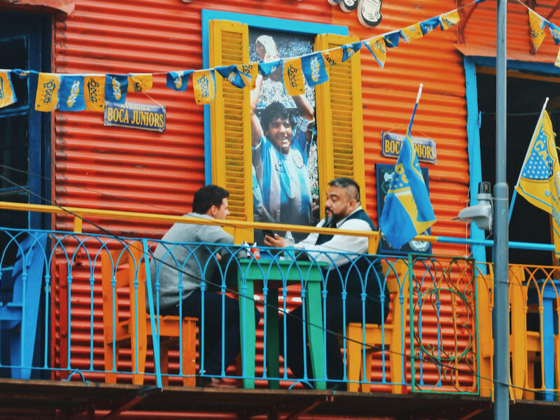 Photo of two men talking while sitting at a table at a bar un Buenos Aires, with a poster of Maradona on the wall behind them.