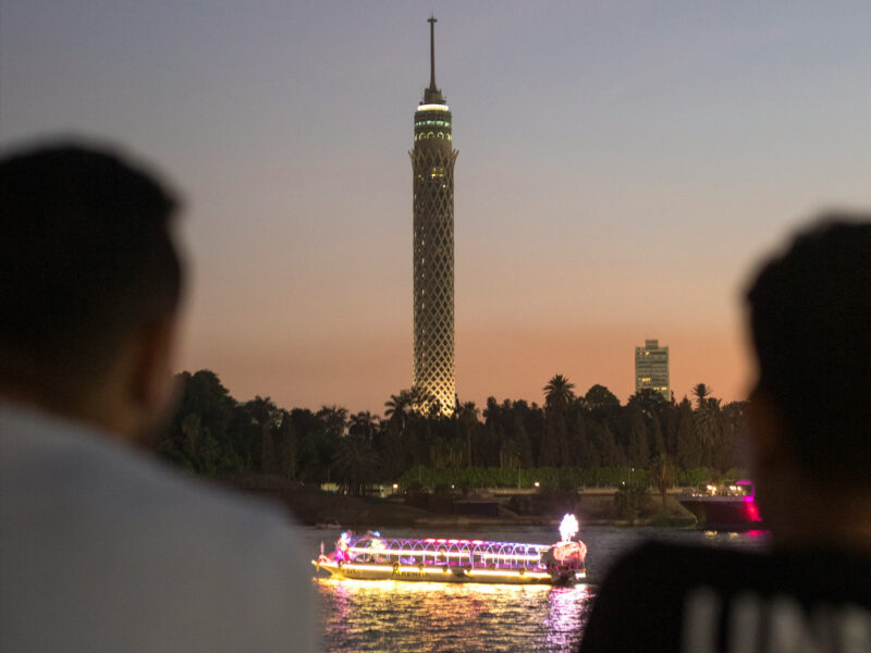 Photo of two men watching boats pass by in Cairo, with a tower in the distance