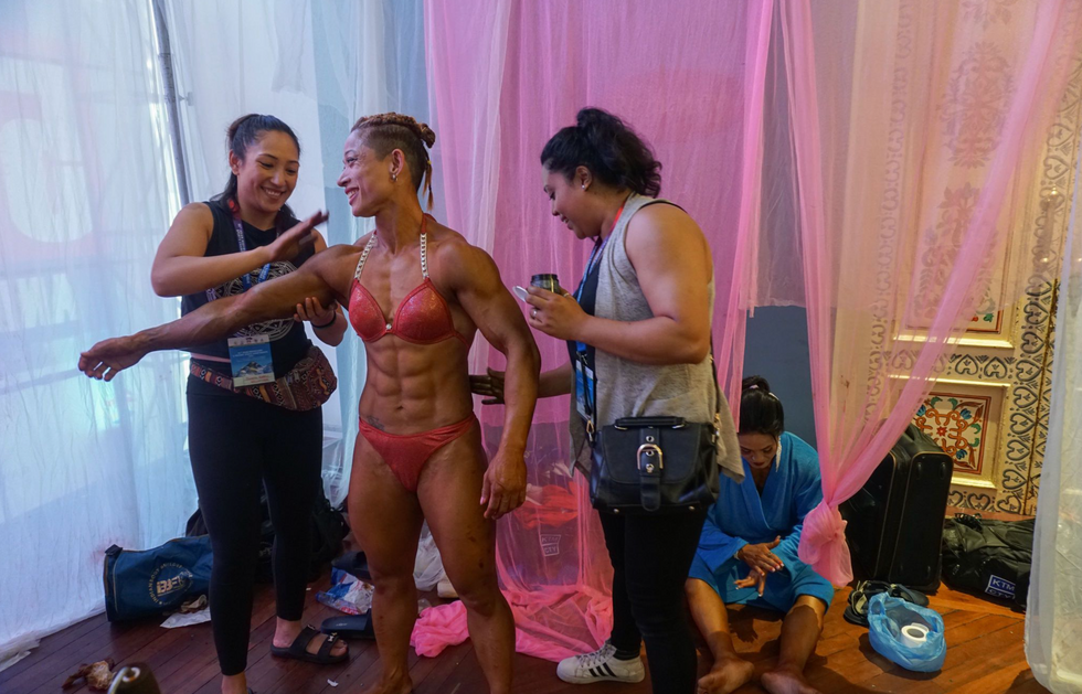Photograph of a female bodybuilder getting ready for a show as women coat her body with a substance.