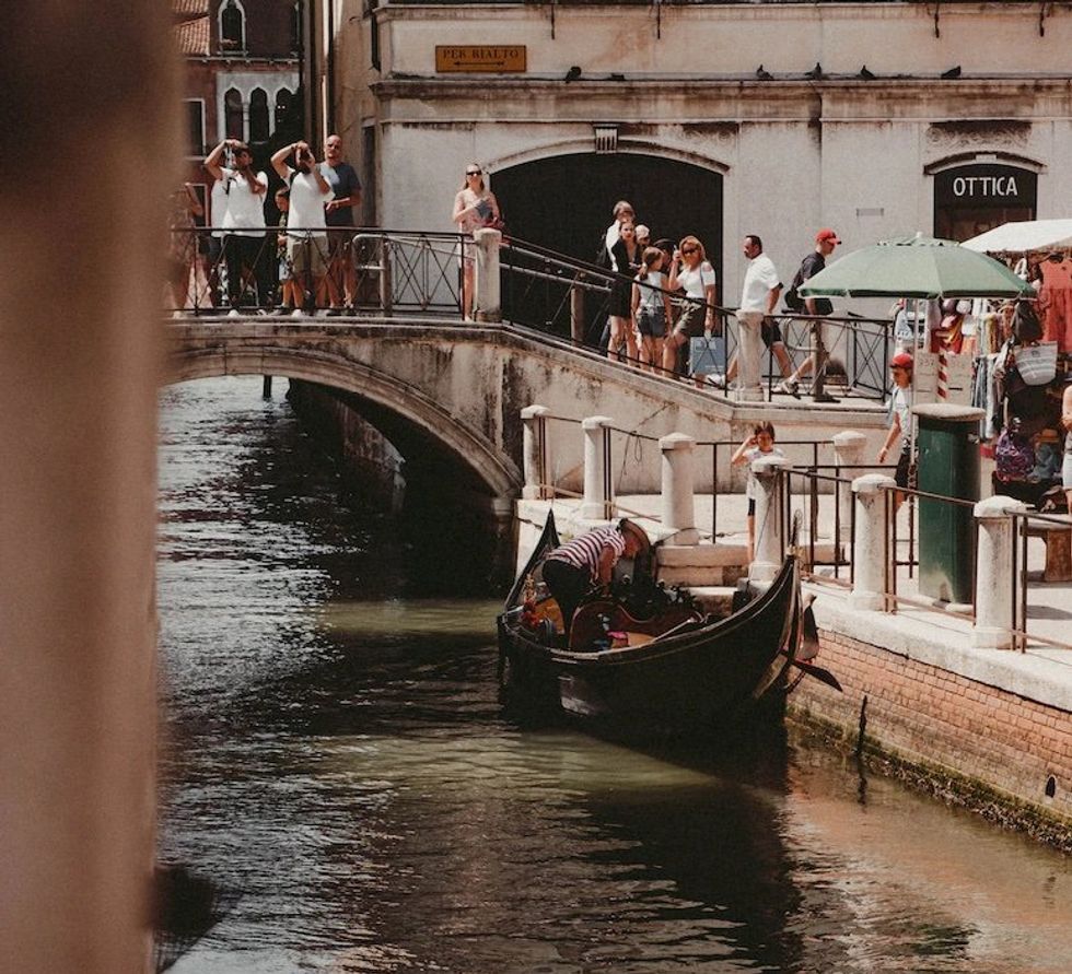 Photograph of a gondola (little Venetian boat) anchored in a canal in Venice as tourists watch, walking over a bridge.